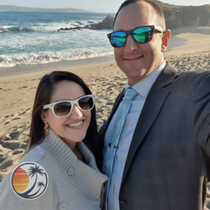 Husband and wife wearing sunglasses and nicely dressed in the foreground. Small semi-transparent Untold Adventures Travel, inc. logo in the lower left corner. Sandy California beach in the background.